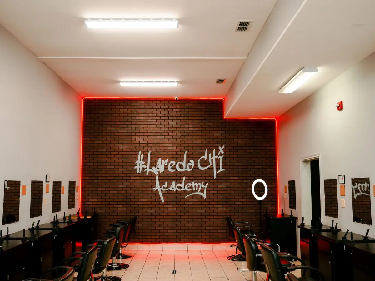 Salon classroom interior with styling stations along both walls and a brick feature wall displaying illuminated “#Laredo CHI Academy” lettering.