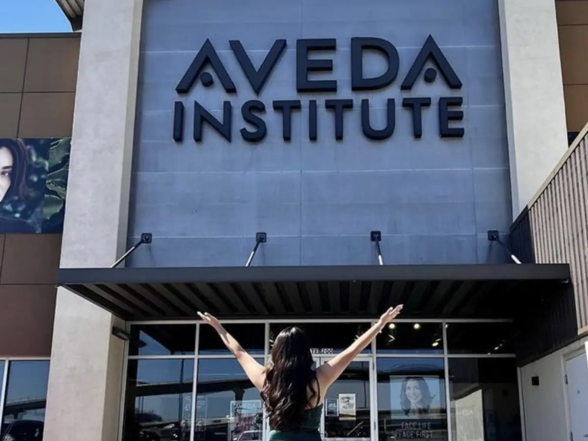 A person with arms raised stands in front of the Aveda Institute entrance beneath large Aveda Institute letters.