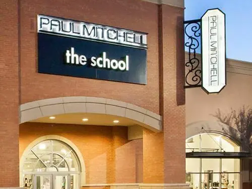 Exterior of a brick Paul Mitchell the school building with large wall-mounted signage, a vertical Paul Mitchell sign, an arched glass entrance, and a storefront window.