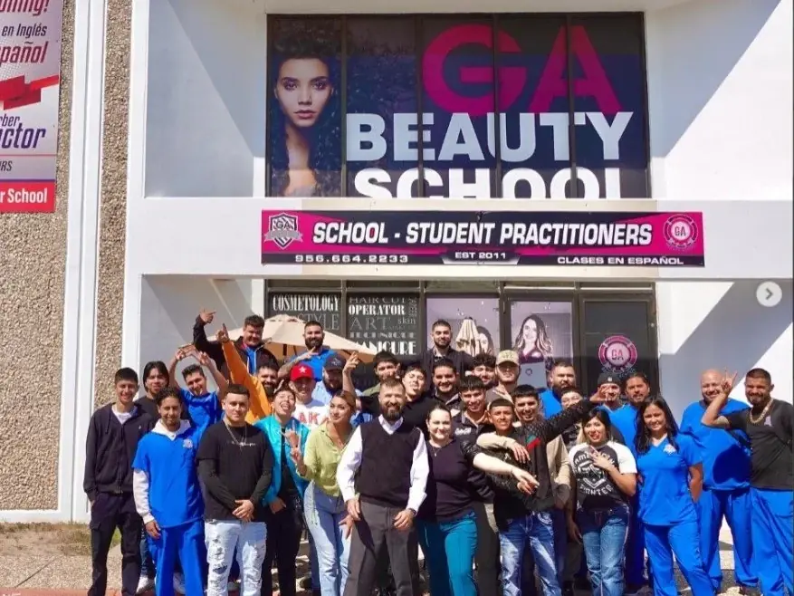 A group of students and staff pose outside the G.A. Beauty School storefront beneath signs reading “GA Beauty,” “School · Student Practitioners,” and “Classes en Español.”
