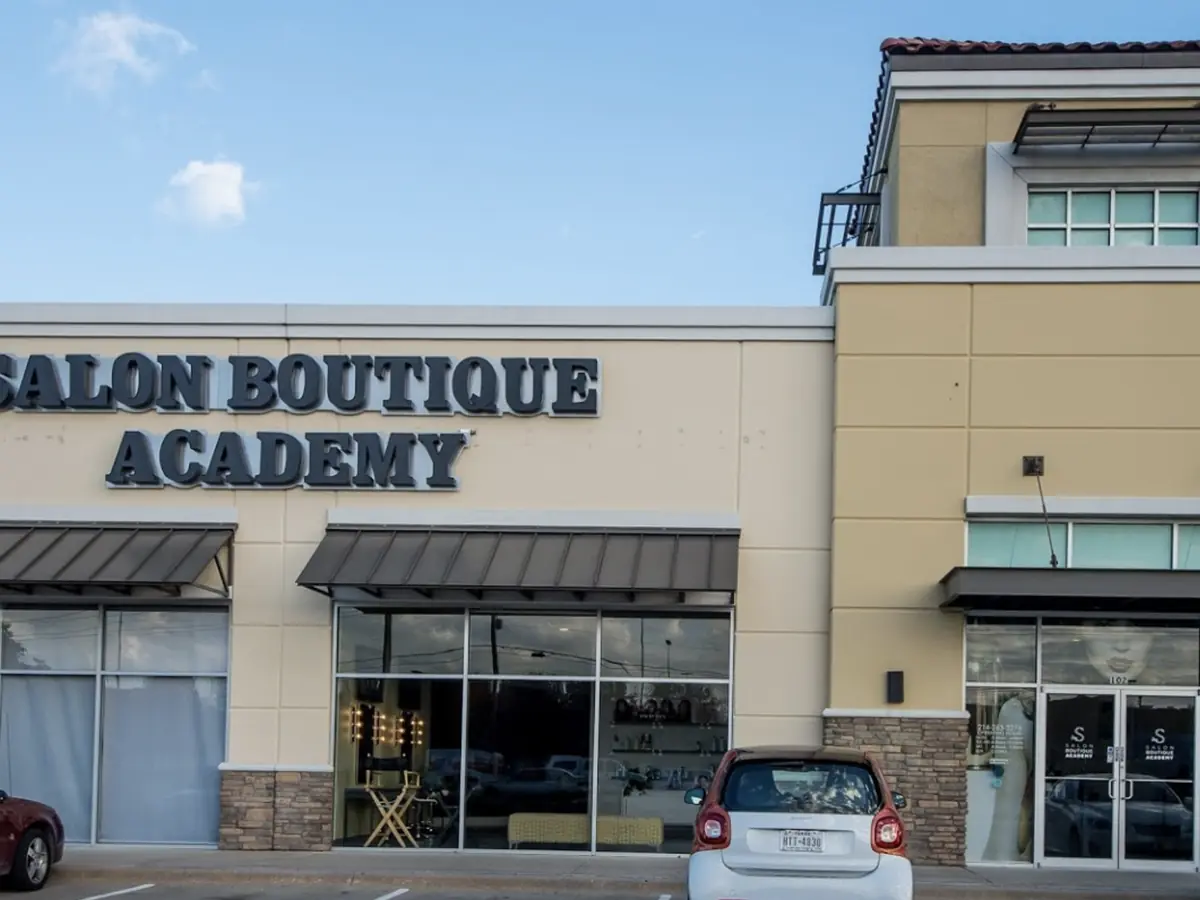 Exterior of Salon Boutique Academy in a beige shopping-center building, with the school name displayed above the storefront entrance and parked cars along the front.