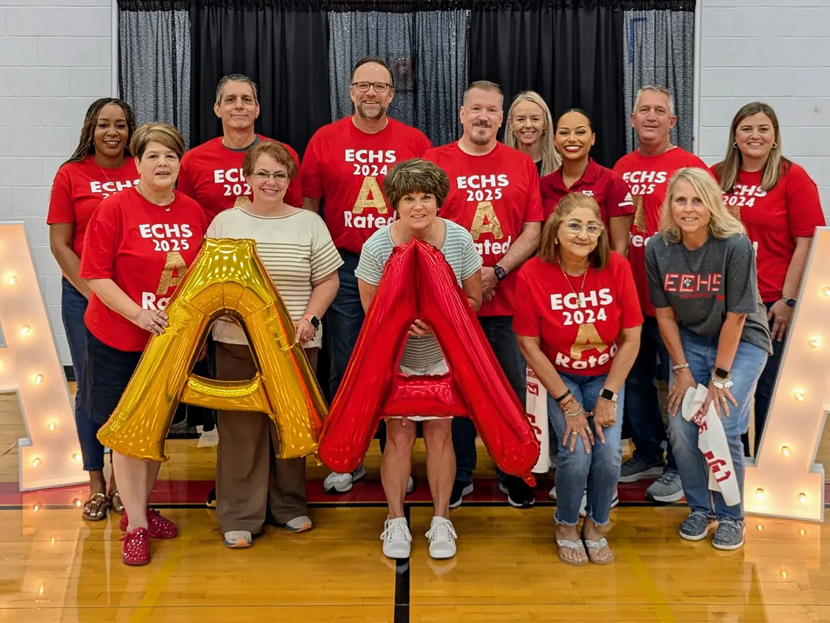 A group of El Campo High School staff and students pose in a gym behind large illuminated letter A decorations and a large gold letter A.