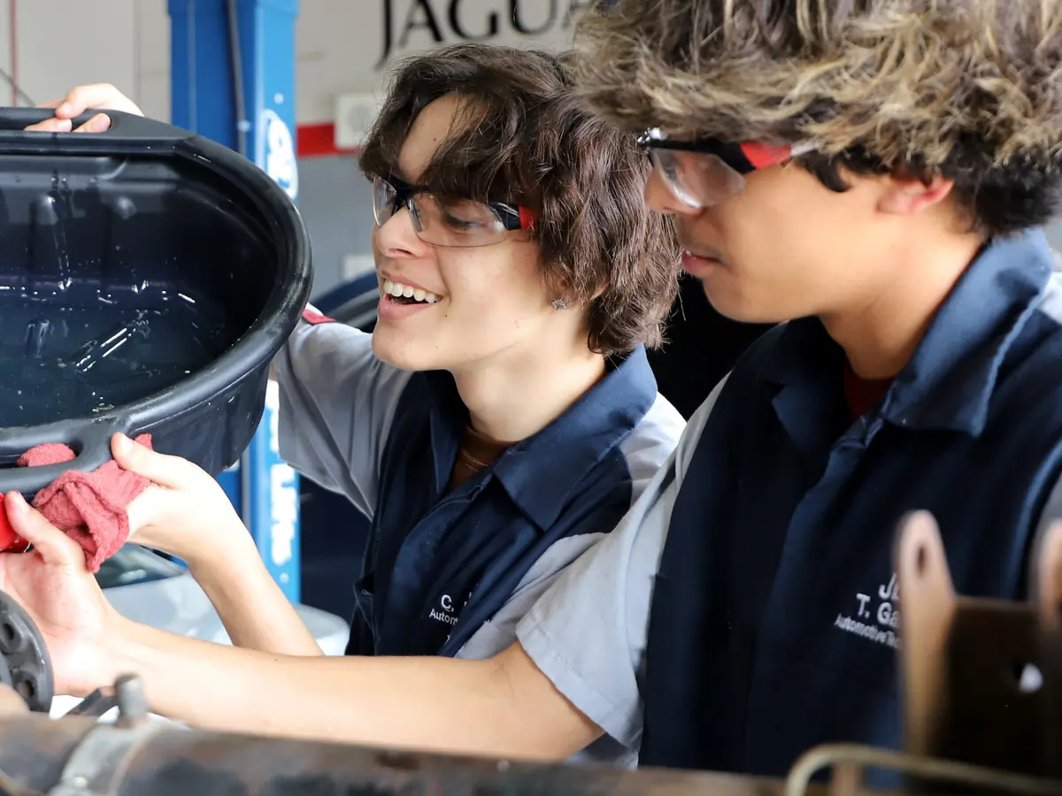 Two students in dark work shirts inspect an automotive part beside red and black shop equipment in a garage.