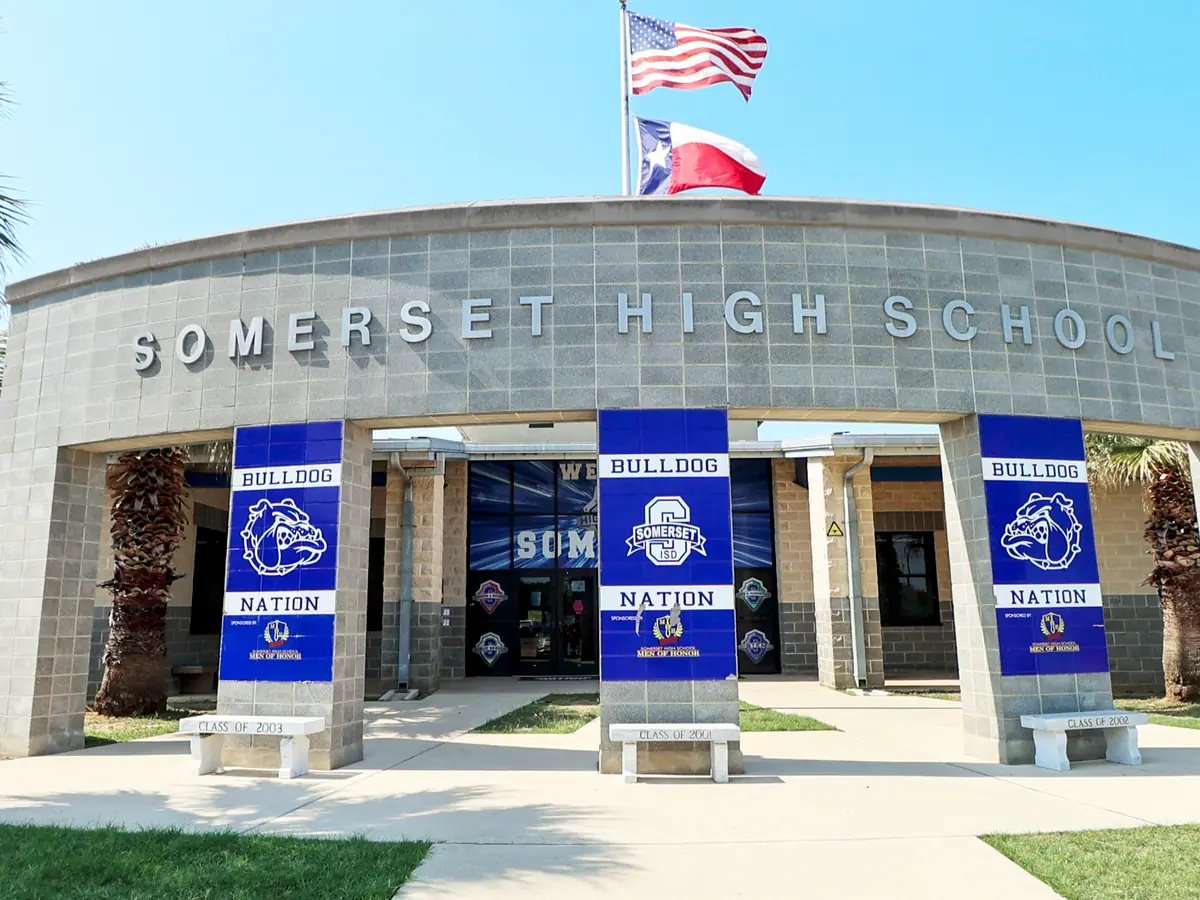 Front entrance of Somerset High School, a curved stone facade with the school name above glass doors, blue banners, flags, palm trees, and accessible parking spaces.