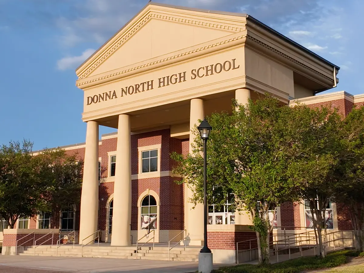 Front entrance of Donna North High School, a red-brick campus building with cream-colored columns, peaked architectural details, trees, and black lamp posts.