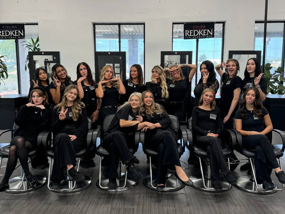 Group of people posing inside a salon in front of a wall sign reading “The Salon Professional Academy,” with salon stations and balloons around them.