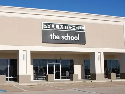 Exterior of a beige commercial building with a prominent “PAUL MITCHELL the school” sign above glass entrance doors and storefront windows.