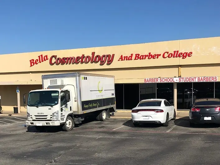 Exterior of Bella Cosmetology And Barber College in a shopping center, with the school name above the entrance and several parked cars in front.