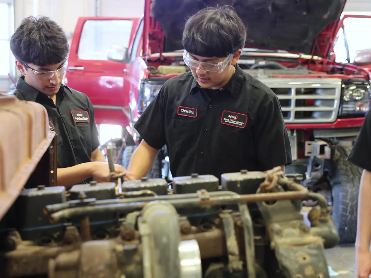 Three students in black automotive-program uniforms work on a vehicle engine in an automotive training lab, with trucks visible behind them.
