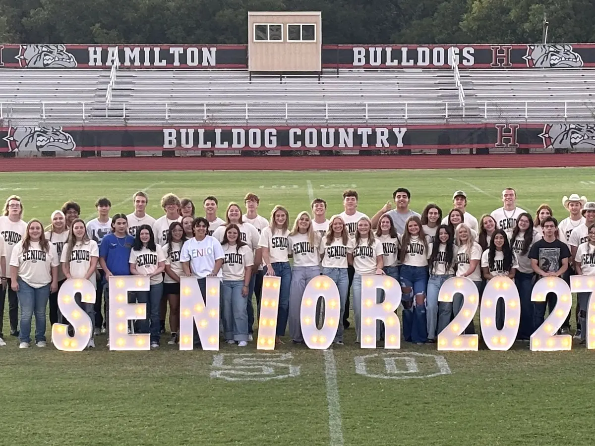A large group of students stands on a football field behind large illuminated “SENIOR 2027” letters; the stadium behind them is labeled “Hamilton Bulldogs” and “Bulldog Country.”