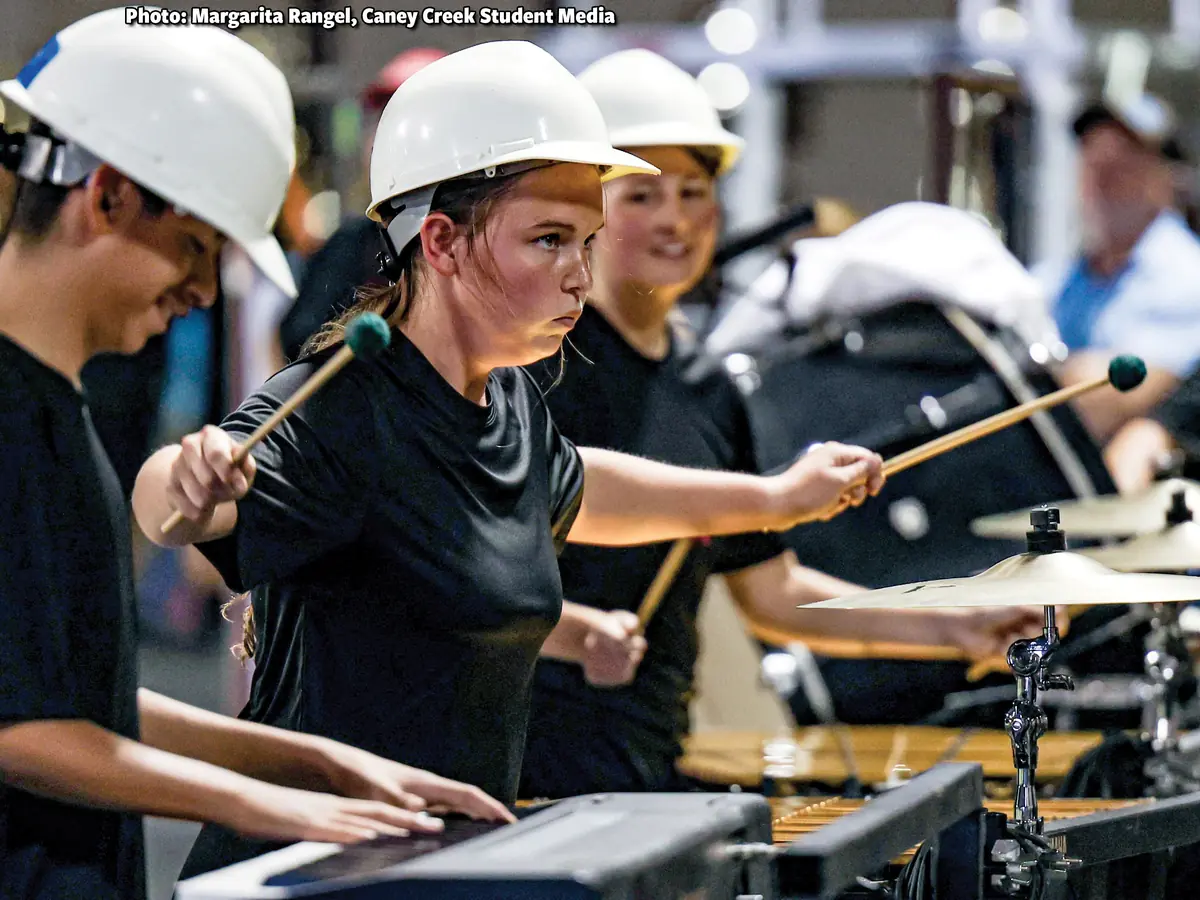 Students wearing white hard hats play percussion instruments, including mallet instruments and drums, in a crowded performance setting.