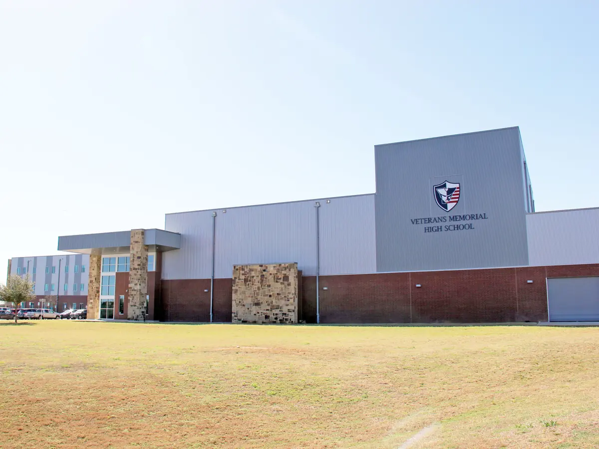 Front exterior of Veterans Memorial High School, a broad gray-and-brick building with a school emblem and name on a tall facade, viewed across a grassy field under a pale blue sky.