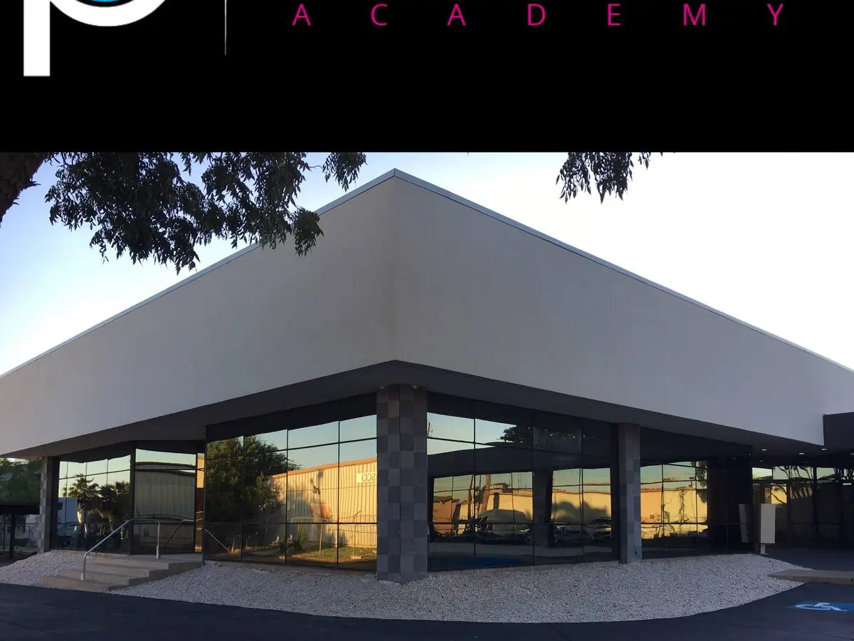 Exterior of The Professional Cosmetology Academy in Laredo, a modern low building with a broad peaked roof, dark stone columns, glass storefront windows, and a covered entrance.