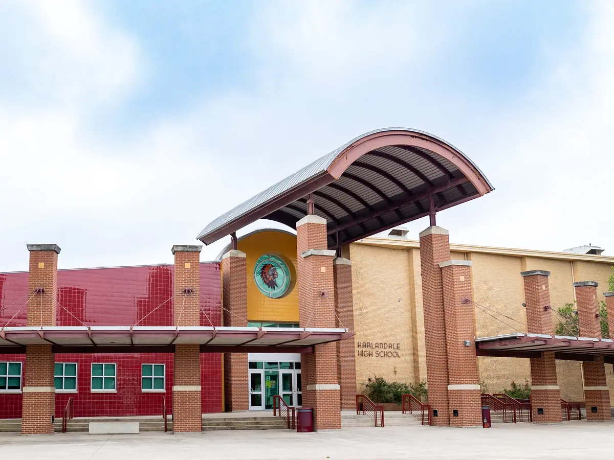 Front exterior of Harlandale High School, a red-brick and tan building with a large curved canopy over the main entrance, school emblem, trees, and windows.