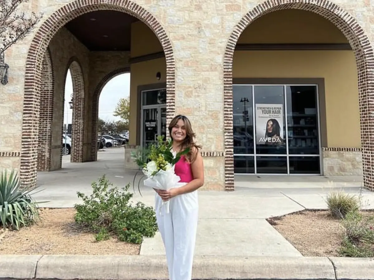 A woman holding a bouquet stands in front of a stone-arched Aveda institute storefront with glass doors and windows.