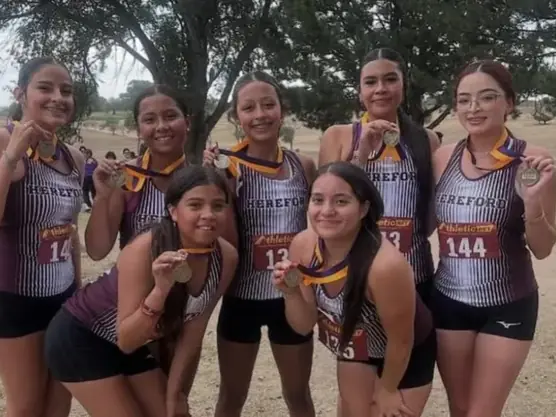 Seven Hereford High School cross-country athletes in maroon-and-white uniforms pose together outdoors beneath trees, with other runners and spectators in the background.