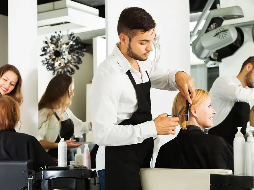 Several stylists in black aprons work with clients seated at salon stations in a bright beauty-school salon, with mirrors, styling tools, and hair dryers visible.
