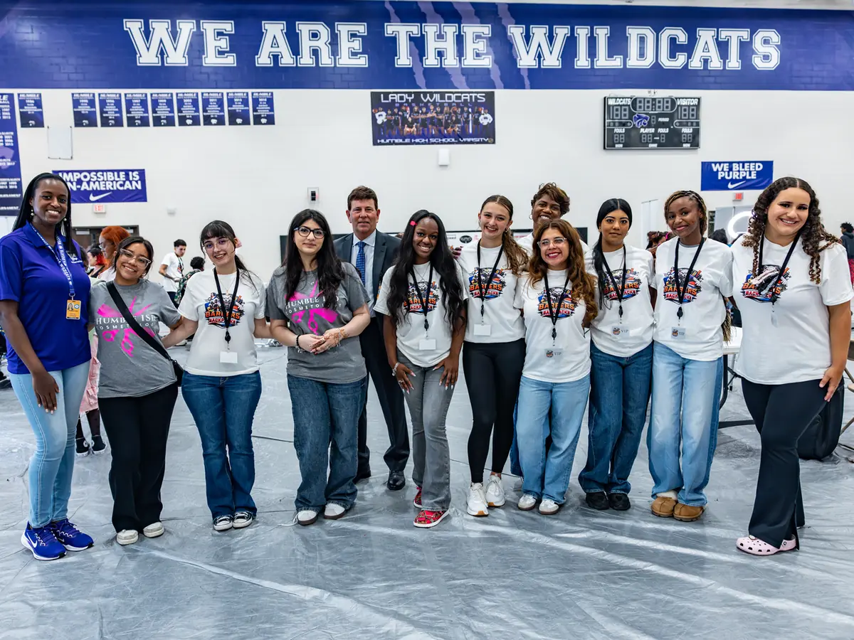 A group of students and an adult staff member stand indoors beneath a large “WE ARE THE WILDCATS” banner, with school display boards behind them.