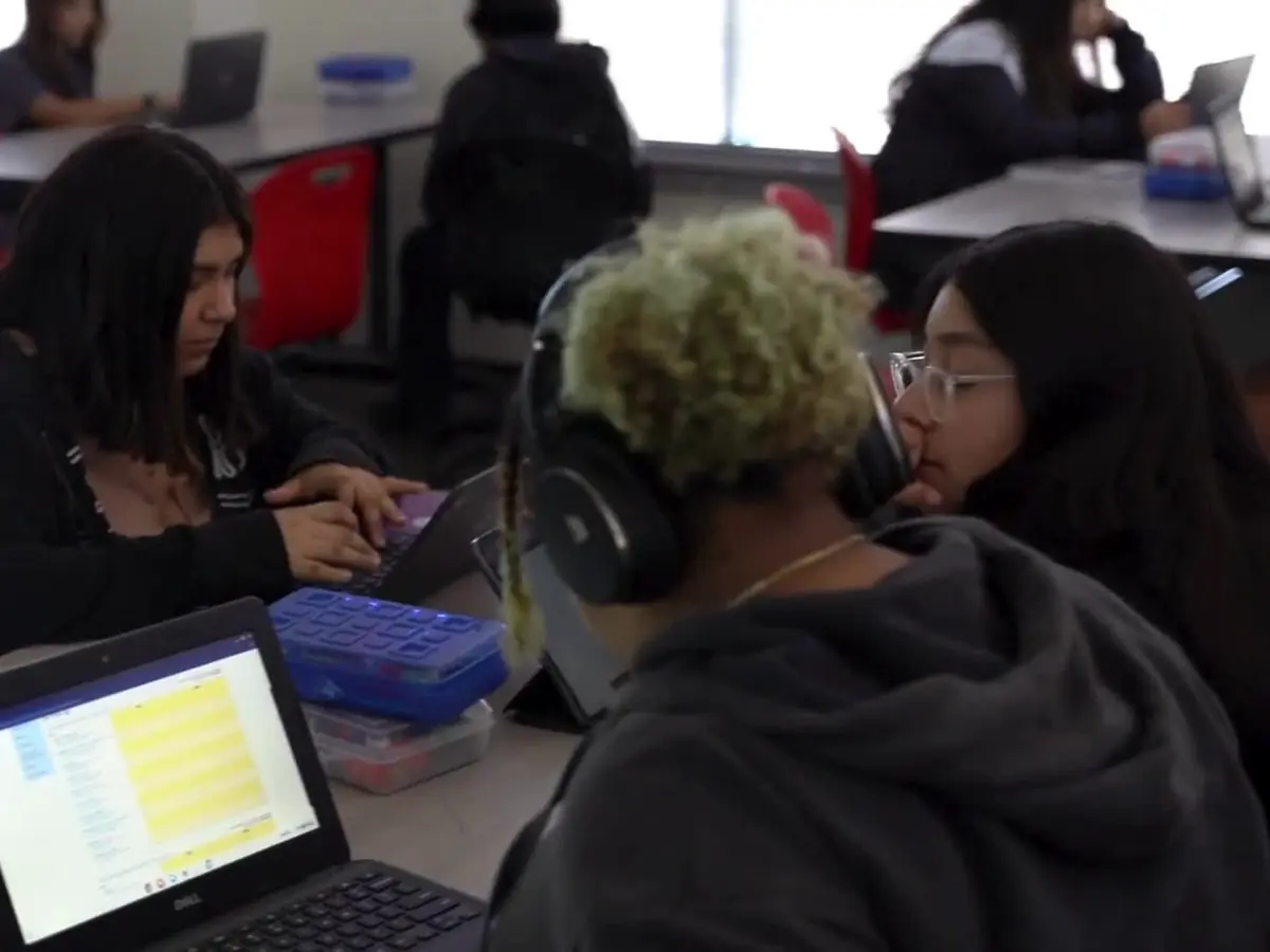 Students work together at tables in a classroom, with laptops, notebooks, and computer monitors visible.