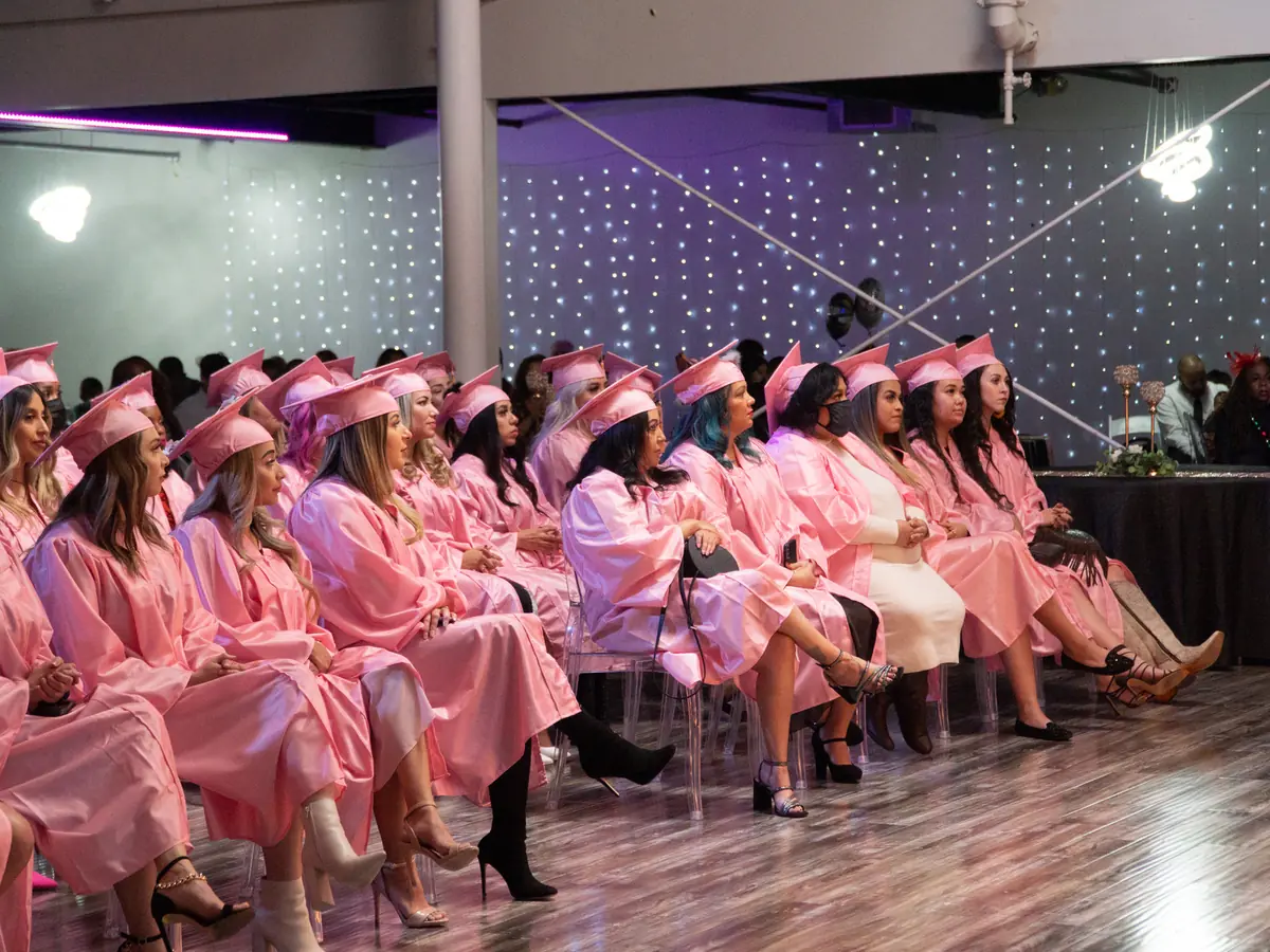 A group of graduates in pink caps and gowns seated in rows inside a dimly lit salon or academy space with purple star-like wall lights.