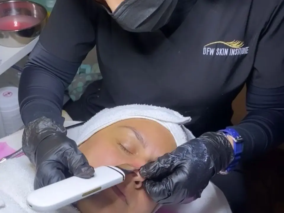 A DFW Skin Institute practitioner wearing gloves uses a handheld device on a reclining client's face during a skincare treatment.