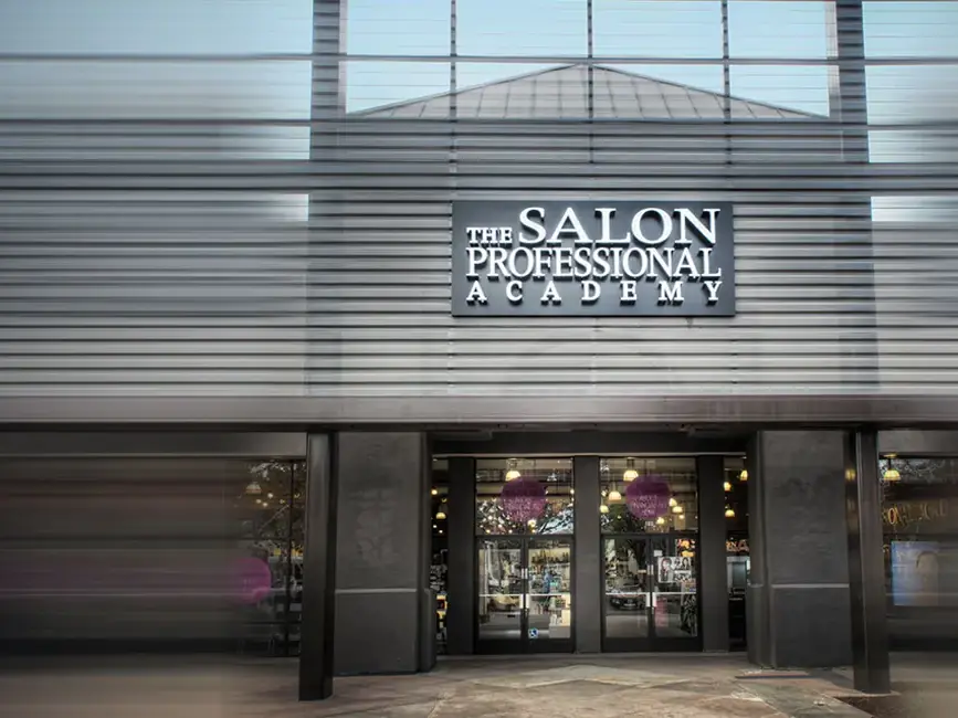 Exterior entrance of The Salon Professional Academy in a modern building, with the academy name displayed above the glass doors.