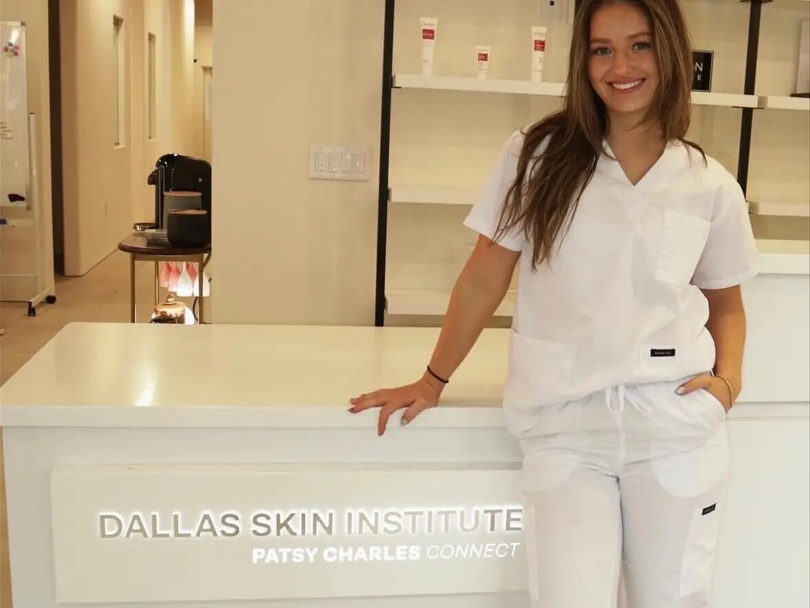 Smiling woman in a white uniform standing beside a reception desk labeled “DALLAS SKIN INSTITUTE PATSY CHARLES CONCEPT,” with product shelves and a treatment room in the background.