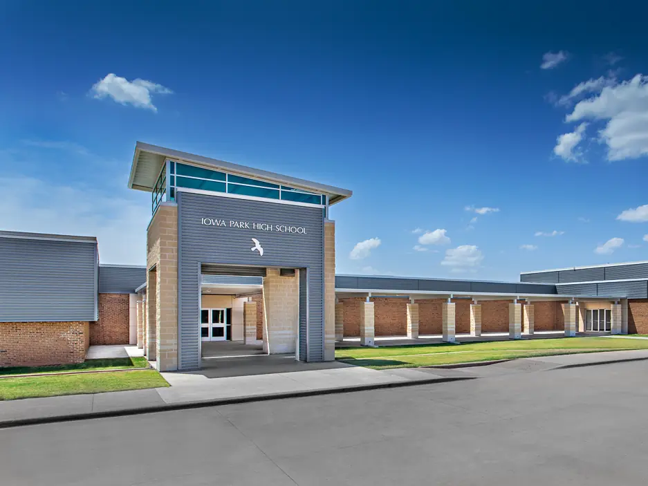 Front exterior of Iowa Park High School, a low brick-and-gray building with a covered entrance tower labeled Iowa Park High School, under a blue sky.