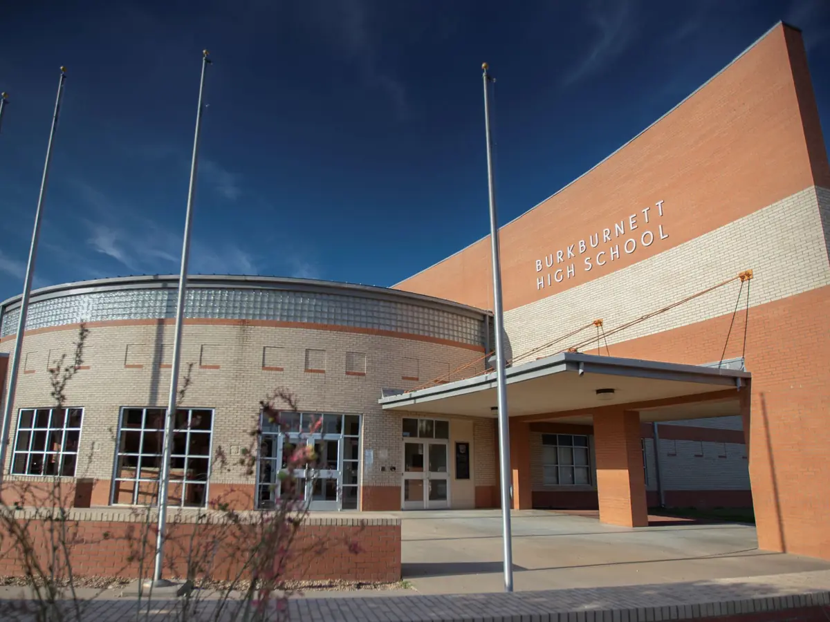 Exterior view of Burkburnett High School, showing the orange-and-white facade, curved glass-front entrance, flagpoles, covered entry, and the school name on the upper wall.
