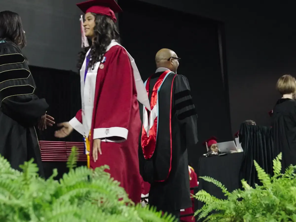 Graduates in caps and gowns on a stage during a James Reese Career and Technical Center ceremony, with ferns in the foreground.