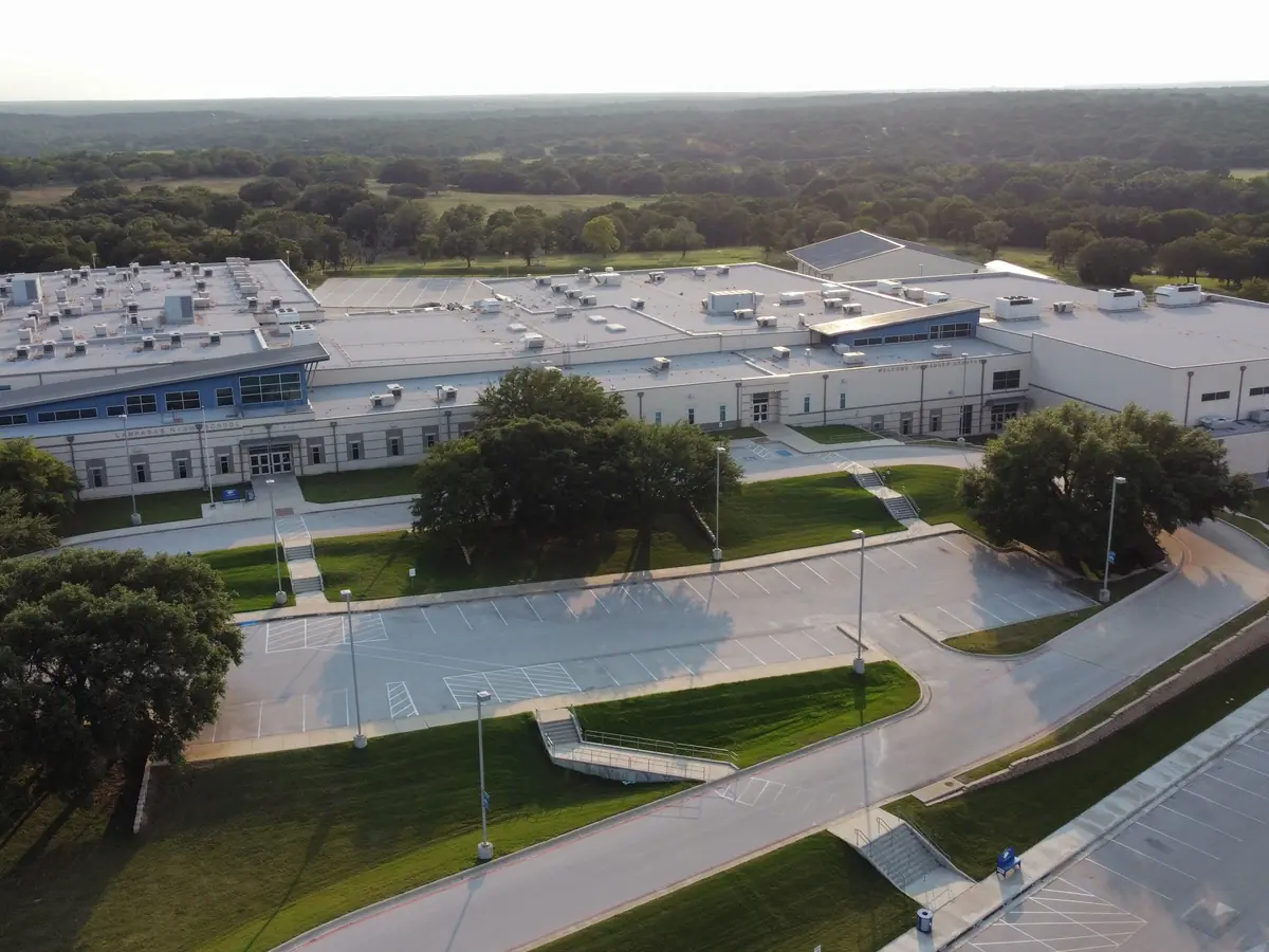 Aerial view of the Lampasas High School campus, showing the main school buildings, paved driveways, parking areas, sports courts, trees, and surrounding landscape.
