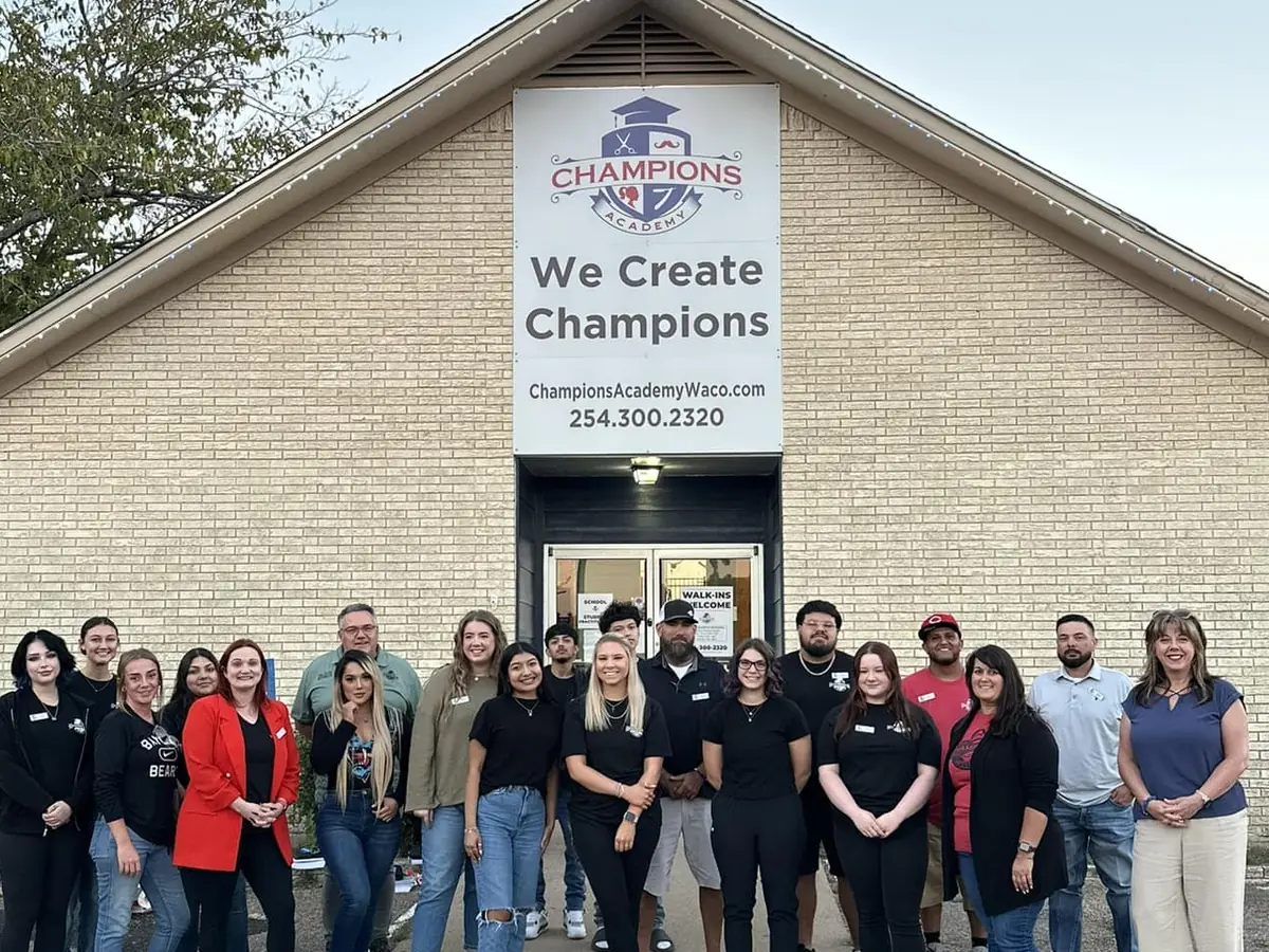 A brick Champions Academy building with a peaked roof and a large sign reading “CHAMPIONS” and “We Create Champions,” with a group of people standing across the entrance and parking area.