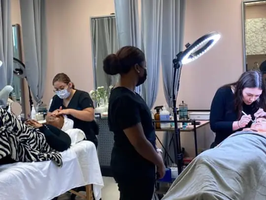 Beauty Kulture Academy training room with several people practicing beauty treatments on clients at treatment beds, separated by gray curtains and lit by a ring light.
