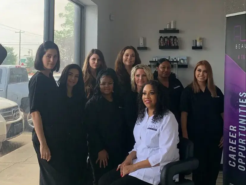 A group of people pose inside a bright beauty academy classroom beside a seated person in a white coat, with salon shelves and a vertical training banner visible.