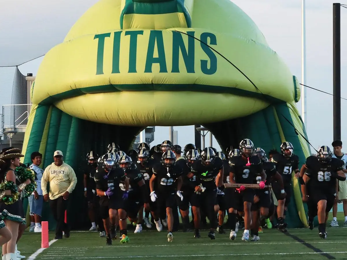 Southwest Legacy Titans football players and cheerleaders emerge from a large green-and-yellow inflatable tunnel onto a football field.
