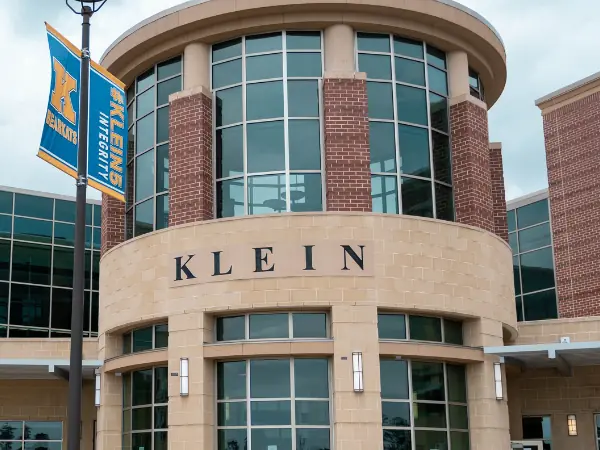 Front entrance of Klein High School, showing a rounded glass-and-brick facade, the word “KLEIN” above the doors, landscaping in front, and a blue school banner on the left.