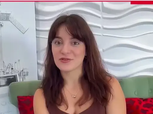 A woman with brown hair sits on a green chair in a beauty-training room, with salon equipment visible on the wall behind her and a white wavy textured backdrop.