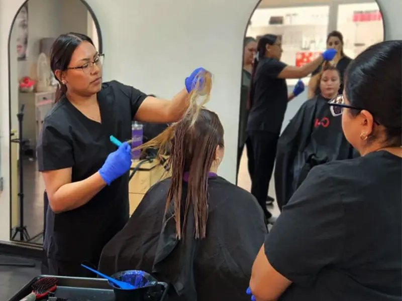 A cosmetology student in black workwear and blue gloves applies hair color to a seated client's wet hair in a salon classroom, with another student nearby and salon mirrors in the background.