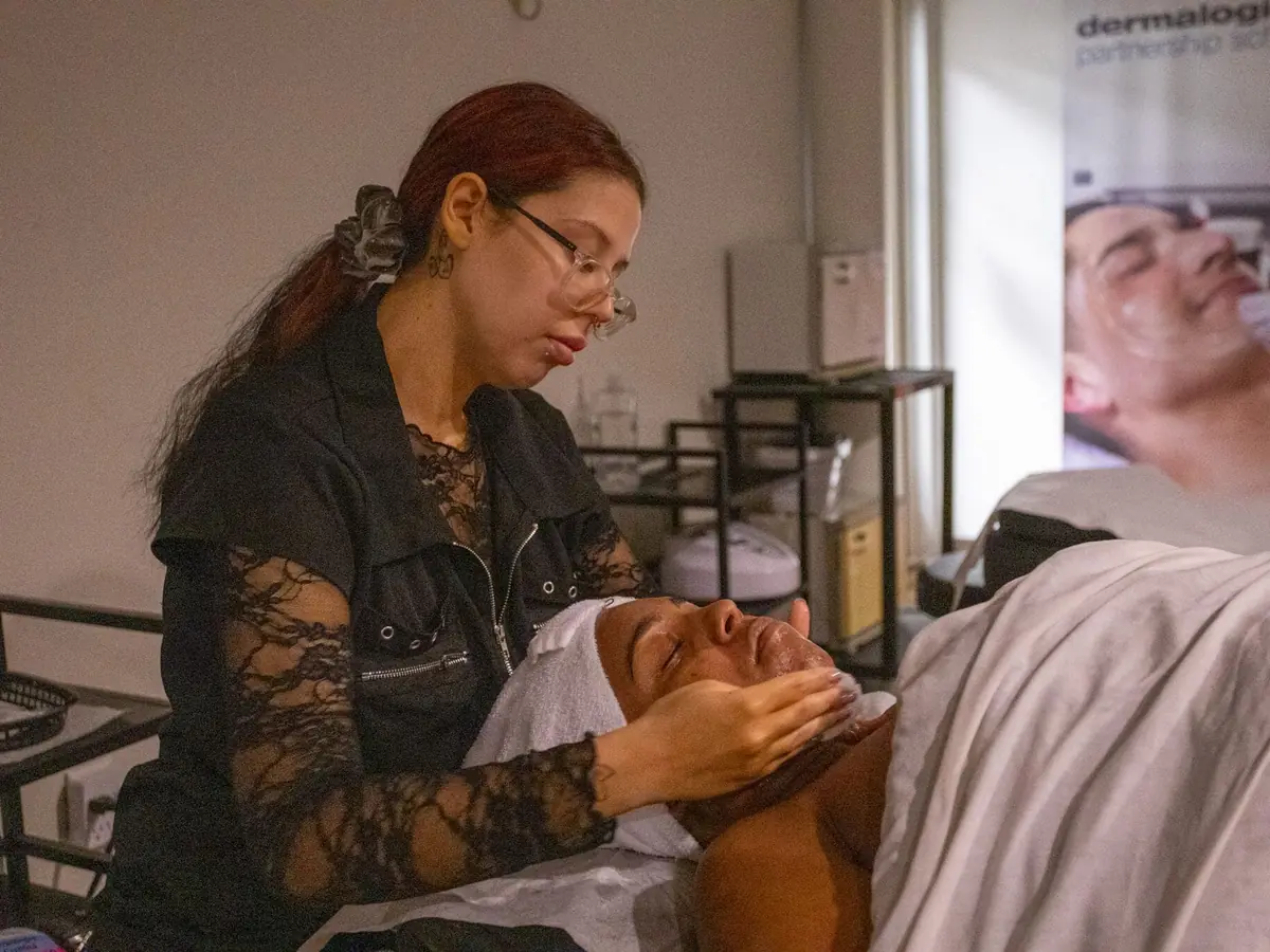 A student in a black uniform massages a reclining client’s face during a facial treatment in a classroom treatment area; another student works in the background.