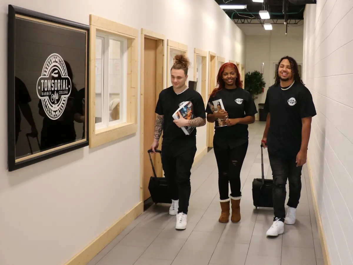 Three students walk down a tiled hallway inside Tonsorial Arts Barber College; a framed Tonsorial Arts logo is visible on the left wall.