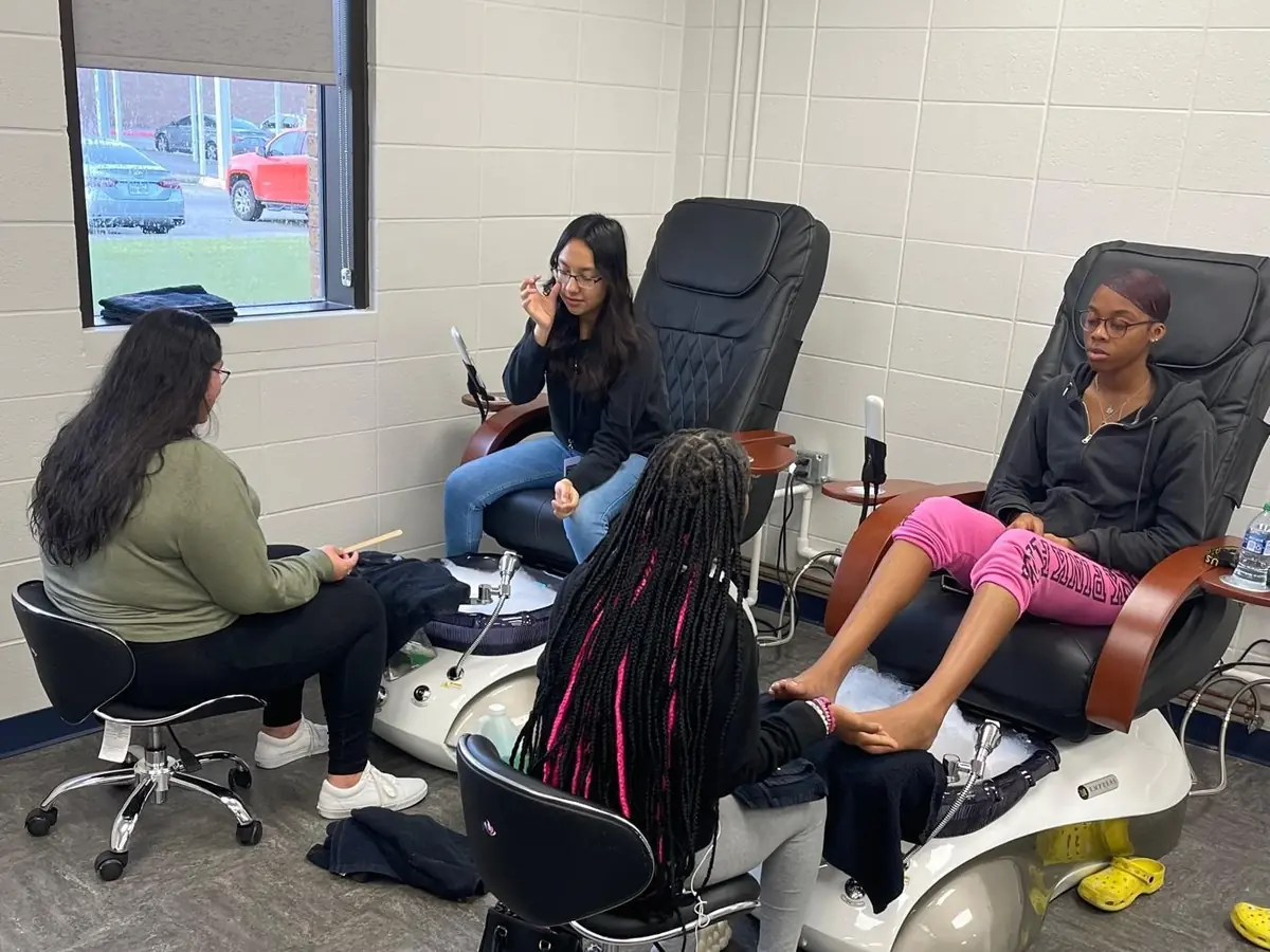 Students and an instructor work in a cosmetology classroom with salon chairs, sinks, and manicure equipment.