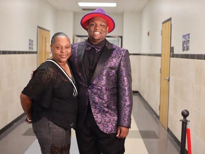 Two people pose together in a tiled school hallway, with classroom doors on both sides and a red velvet rope beside them.