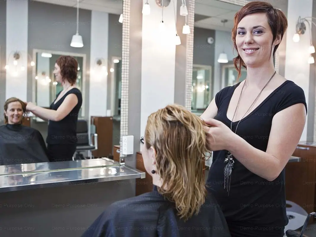 A stylist stands beside a seated client in a salon, holding the client's hair in front of a mirrored styling station with pendant lights overhead.