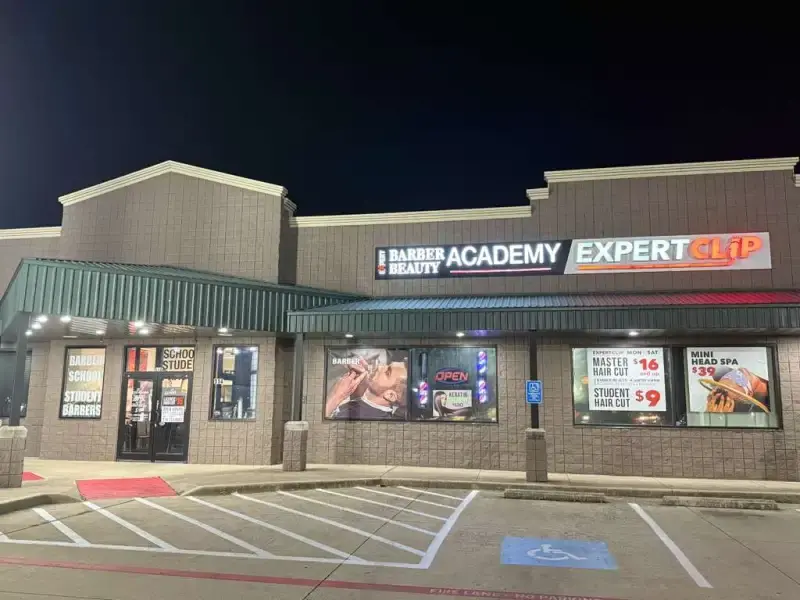 Nighttime exterior of the Expert Barber Beauty Academy storefront in a strip center, with illuminated academy signage, barber and beauty advertisements in the windows, and marked parking spaces in front.