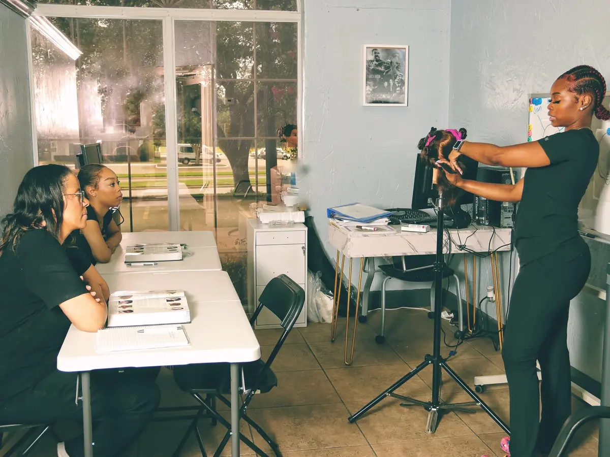 Instructor demonstrates hairstyling to seated students in a classroom with tables, mirrors, and a large window.