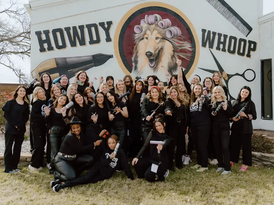A large group of people in black clothing pose on the lawn in front of a white building painted with a colorful mural featuring the words “HOWDY” and “WHOOP,” a blow dryer, scissors, and a stylized dog.