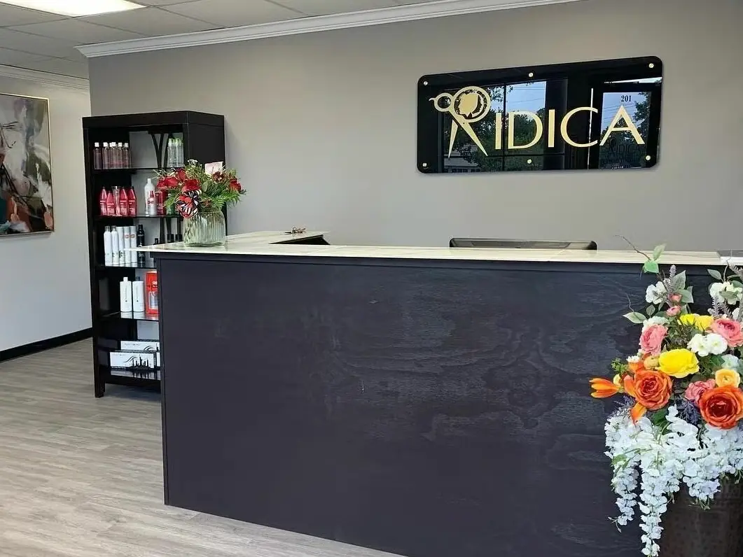 Salon reception desk with a black RIDICA sign on the wall, product shelves, framed artwork, and a colorful flower arrangement.