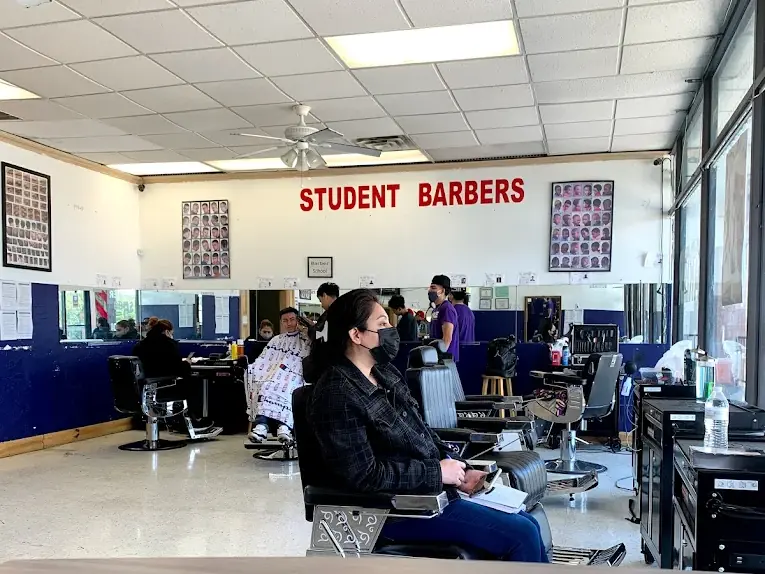 Interior of a barber classroom with multiple barber chairs, students and instructors, workstations, and a wall sign reading “STUDENT BARBERS.”