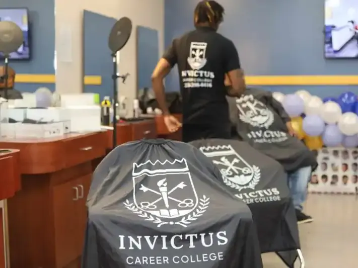 Inside a cosmetology training salon, several black capes display the words “INVICTUS CAREER COLLEGE,” with styling stations, mirrors, and balloons in the background.