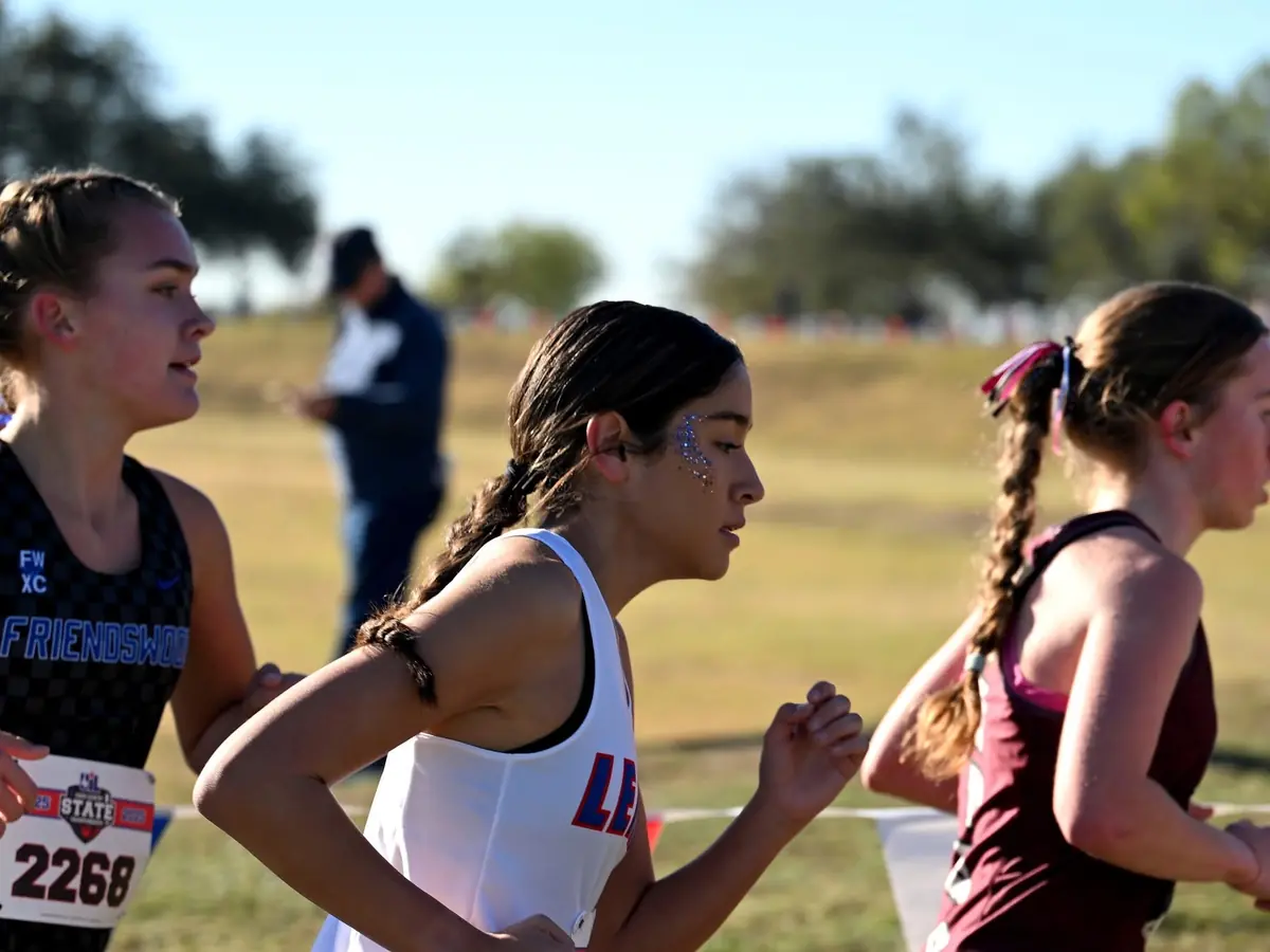 Three teenage runners compete across a grassy field; the center runner wears a white uniform with blue and red LHS lettering.